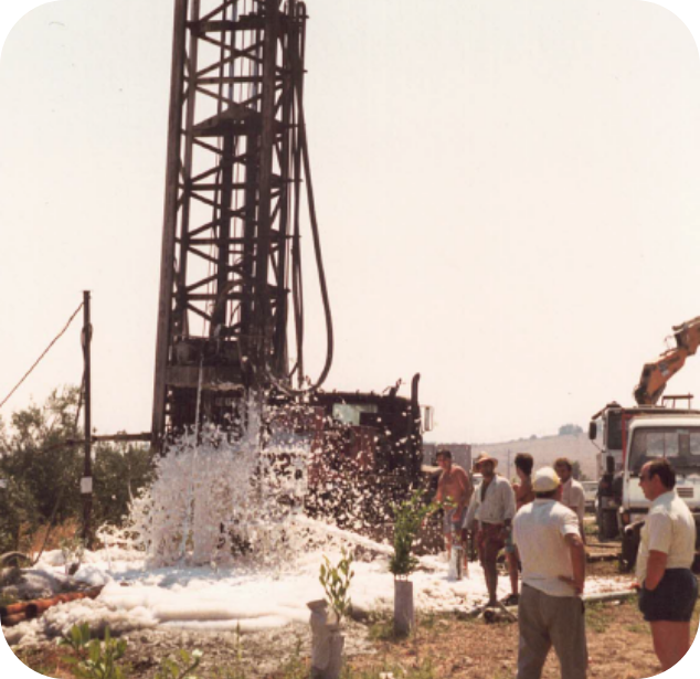 Foto storica dello scavo di un pozzo con getto d’acqua e persone al lavoro nei campi.
